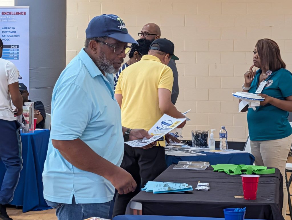 A man in a blue shirt and cap looks at papers while standing at a table during an indoor event with other people.