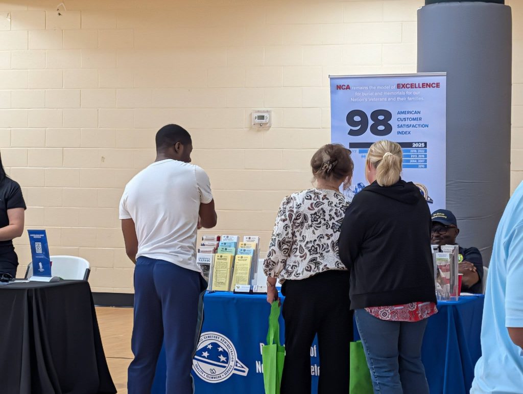 Three people stand at an informational booth with pamphlets and a sign displaying "98". The booth has a blue table cover and is indoors.