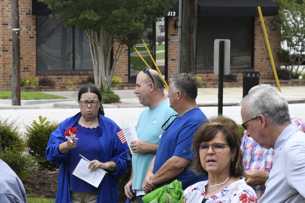A group of adults stand outdoors having a conversation, some holding papers, with trees and buildings in the background.