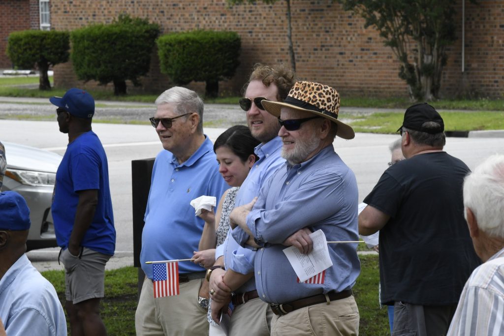 A group of people standing outdoors, some holding small American flags, dressed casually. One person wears a leopard-print hat. A brick wall and bushes are in the background.