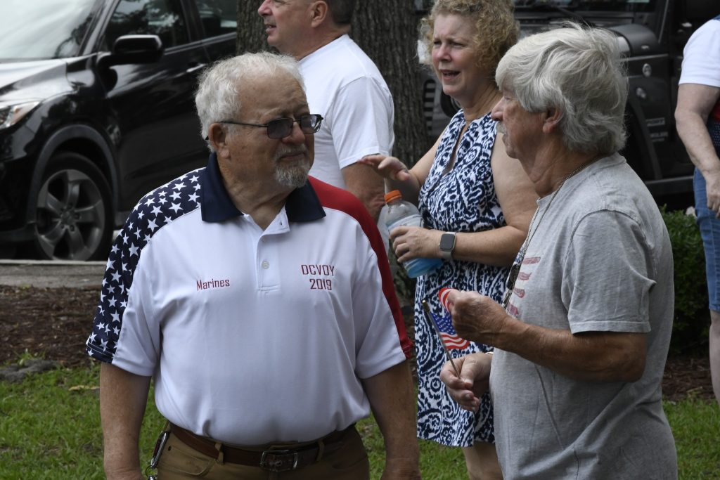 Older adults socializing outdoors, one man wearing a patriotic shirt, others chatting and smiling, with cars and trees in the background.