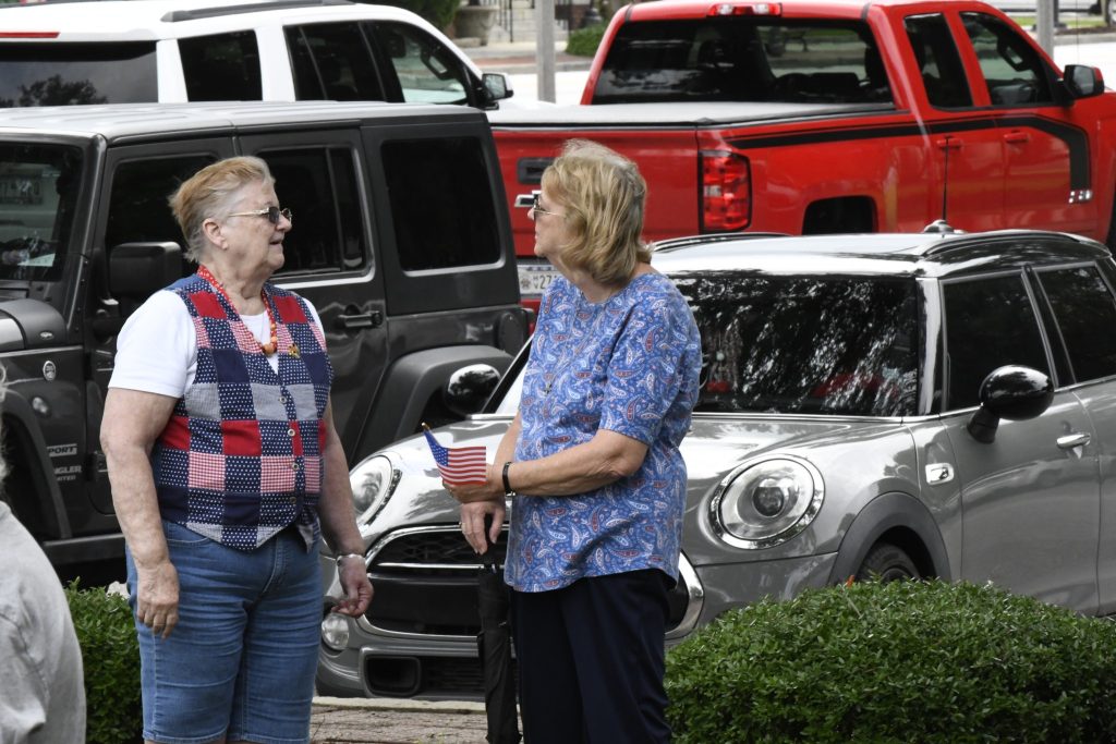 Two women standing and talking in a parking lot, with parked cars, including a Mini Cooper and a red truck, visible in the background.