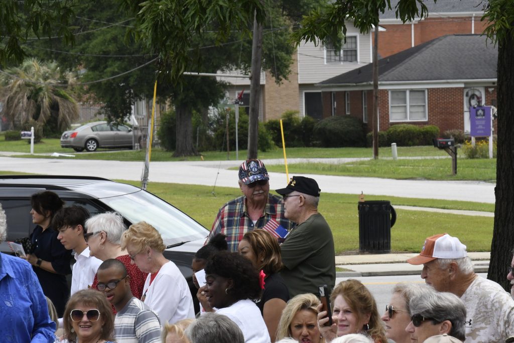 A group of people gathered outdoors, with two veterans wearing military caps standing among the crowd in a residential neighborhood.
