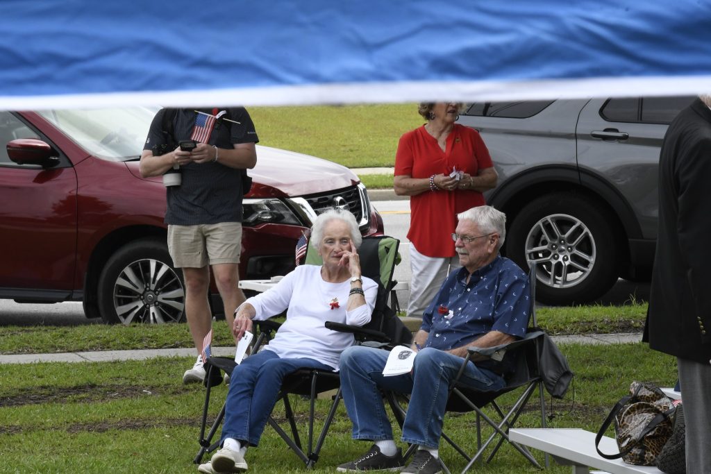 Two elderly people sit on lawn chairs under a canopy, with three people standing behind them near parked cars on a grassy area.