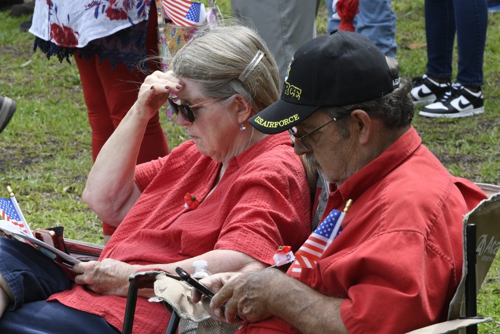 older man and woman sitting outdoors, both wearing red shirts. The man wears a veteran cap and holds a small American flag.