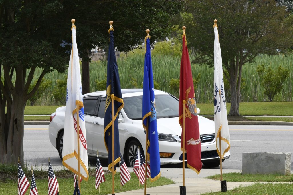 White car parked on street behind six military flags and several small American flags on a grassy area.