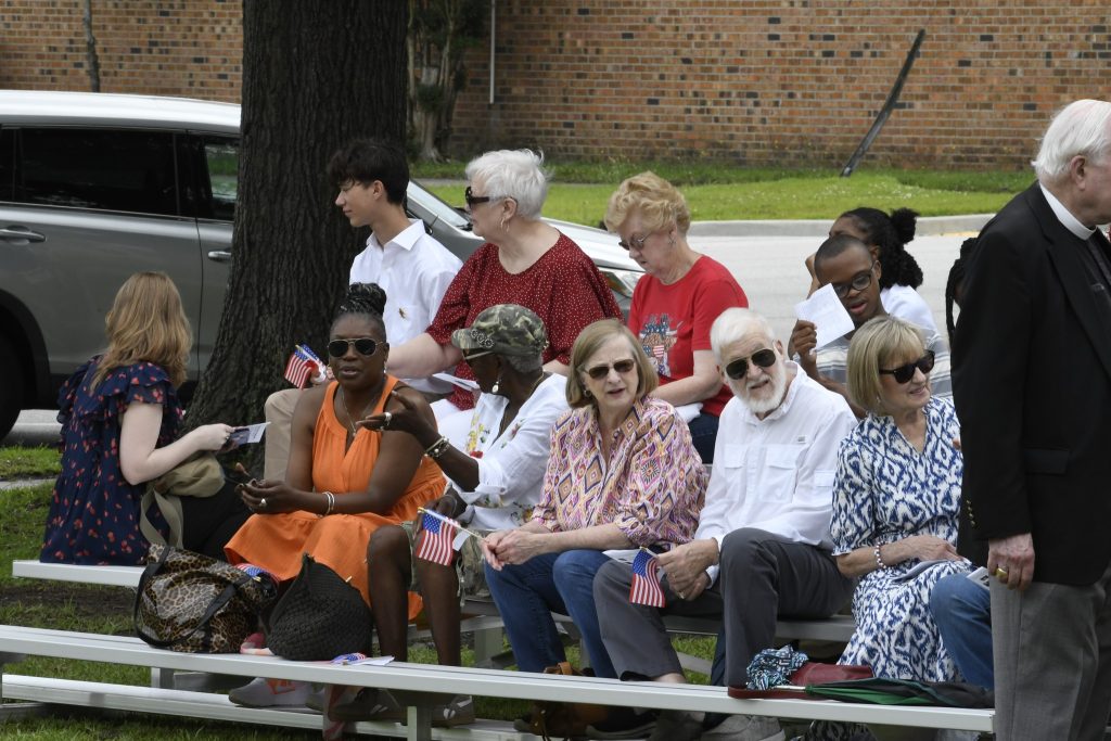 A group of people sitting on bleachers outdoors, holding small flags, watching an event, with trees and a brick wall in the background.