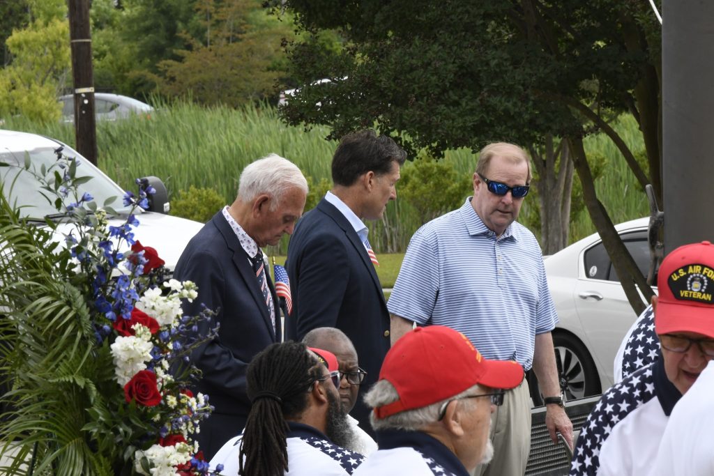 Several men, some in suits and some casually dressed, gather outdoors near a floral arrangement, with trees and parked cars in the background.