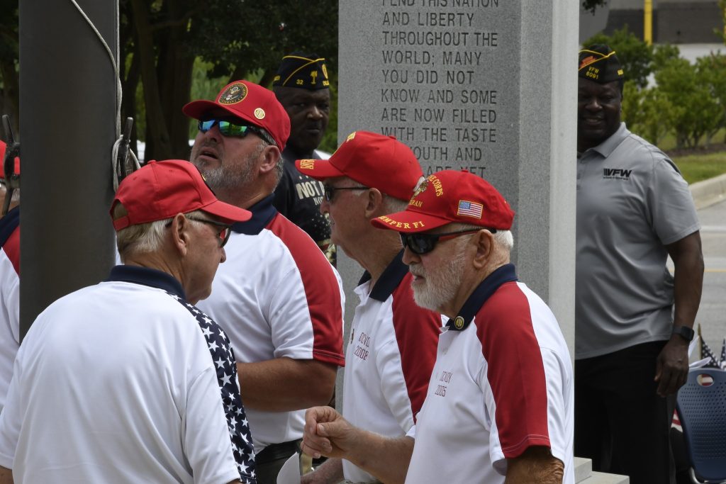 A group of older men wearing red hats and white shirts stand together outdoors near a memorial stone engraved with text.