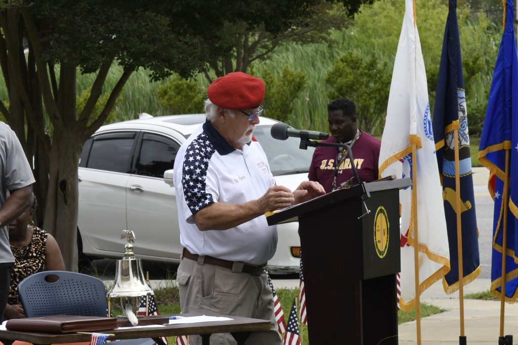 older man wearing a red beret and patriotic shirt speaks at an outdoor podium with flags, people, and cars in the background.