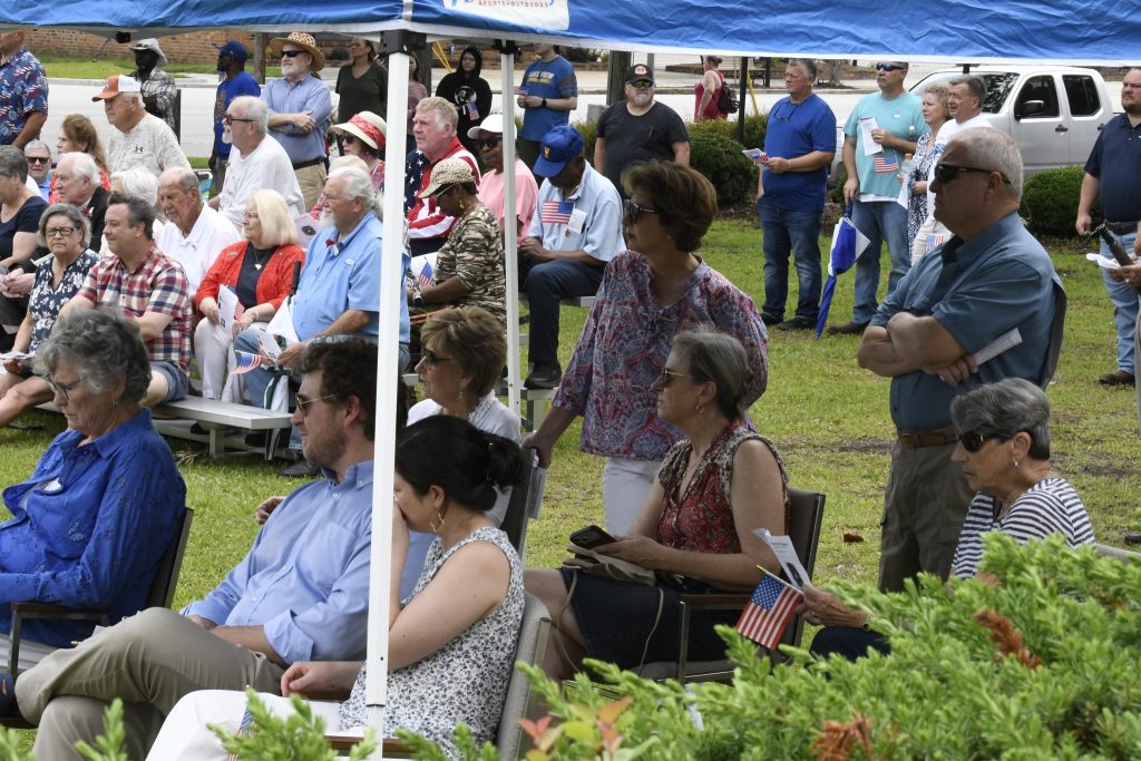 People sitting and standing under a tent at an outdoor event, some holding small American flags, with more people gathered in the background.