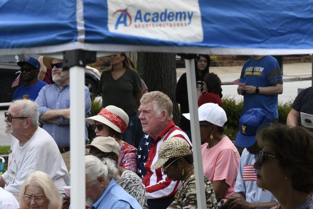 People seated under a blue Academy Sports tent at an outdoor event, with some wearing hats and festive attire, and others standing in the background.