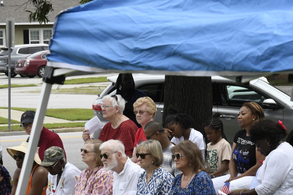 A group of people of various ages sit outdoors under a blue canopy, watching an event on a sunny day.