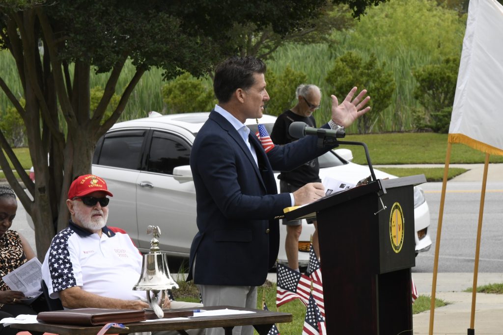 A man in a suit speaks at an outdoor podium with American flags, while another man in a red cap sits nearby.