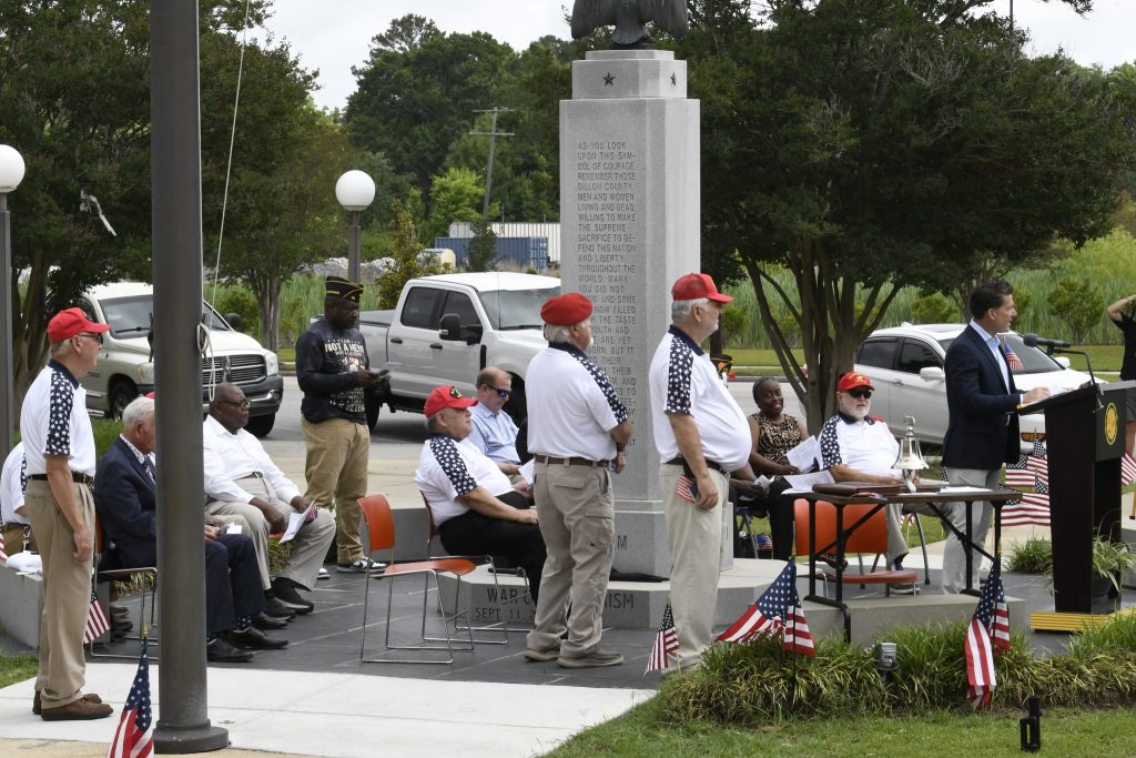 People gathered outdoors at a memorial site, some seated and others standing, with a monument in the background and American flags displayed nearby.