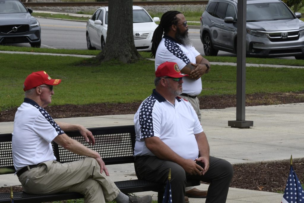 Three men in matching white shirts with star patterns, two sitting on a bench and one standing, outdoors near parked cars and trees.
