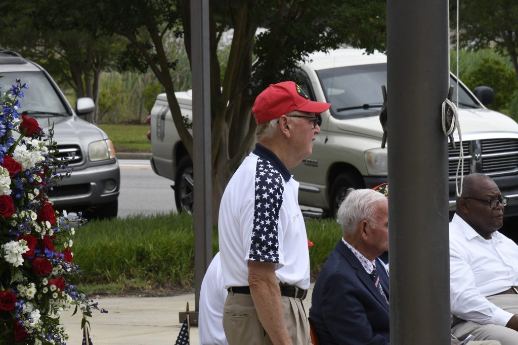 older man in a red cap and patriotic shirt stands outdoors near another seated man in a suit, with cars and greenery in the background.