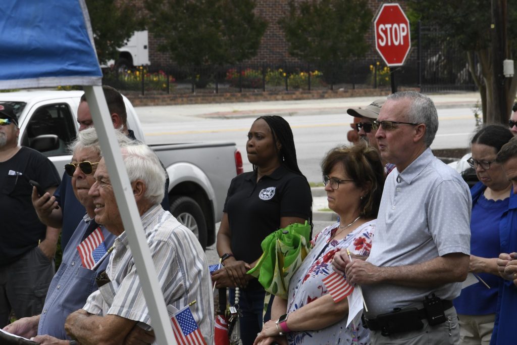 A group of people stand outdoors near a street, some holding small American flags, with a stop sign and parked vehicles visible in the background.