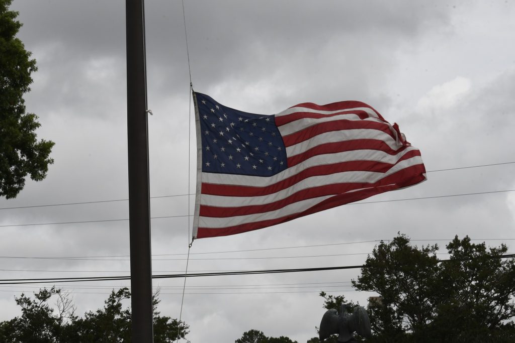 The American flag is flying at half-mast on a cloudy day with trees and power lines in the background.