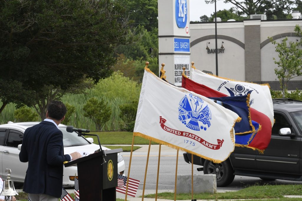 A person speaks at an outdoor podium beside several military flags; a building and sign are visible in the background.