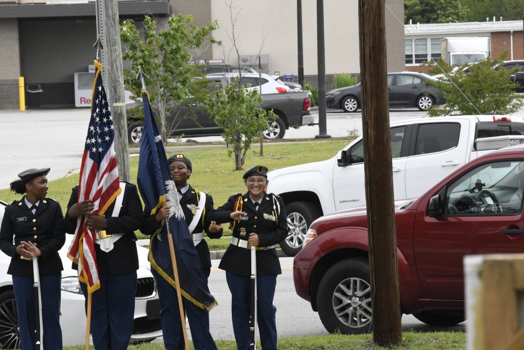 Three uniformed people stand outside holding flags beside parked cars and a pole, appearing to participate in a formal ceremony or parade.