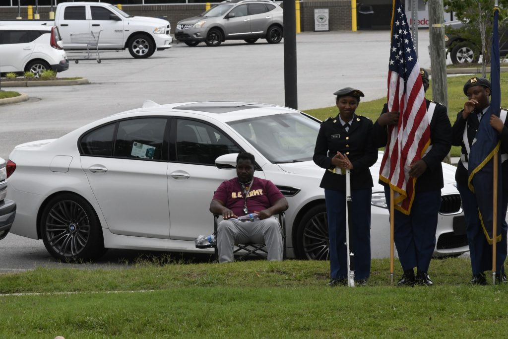 Two uniformed guards stand with the American flag on a grassy area, while a seated man watches in front of a parked white car.