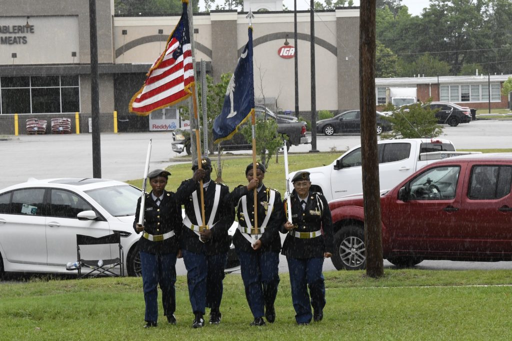 Four uniformed military personnel carry flags, including the American flag, while marching on grass with parked cars and a building in the background.