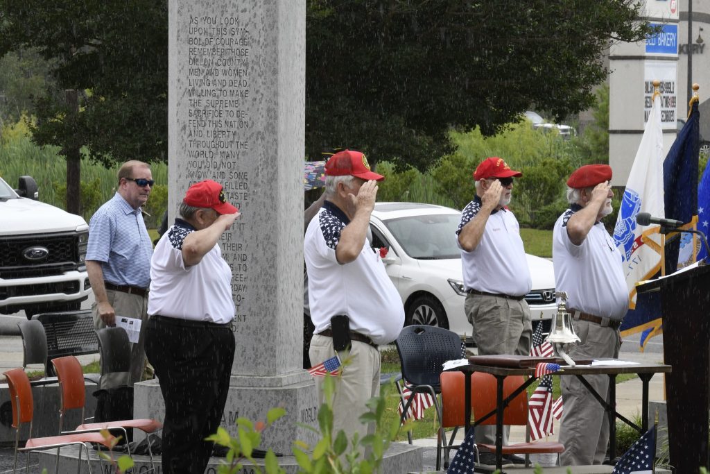 Several people in matching shirts and red hats salute in front of a memorial monument, with American flags and a vehicle visible in the background.
