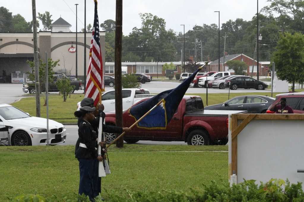 Two uniformed individuals stand outdoors holding American and blue flags near a sign, with parked cars and buildings in the background on a cloudy day.