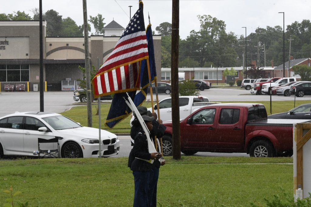 A uniformed individual holds multiple flags, including the American flag, outside on a grassy area with parked cars and buildings in the background.