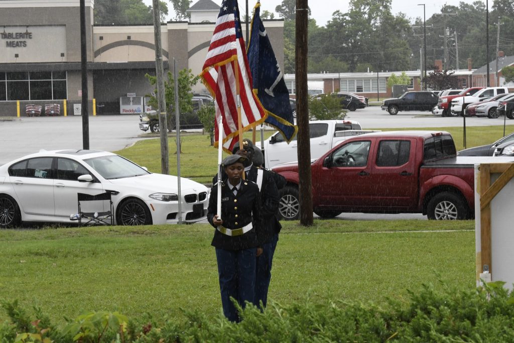 A uniformed honor guard carries the American flag and military flags outdoors, with cars and buildings visible in the background on a cloudy day.