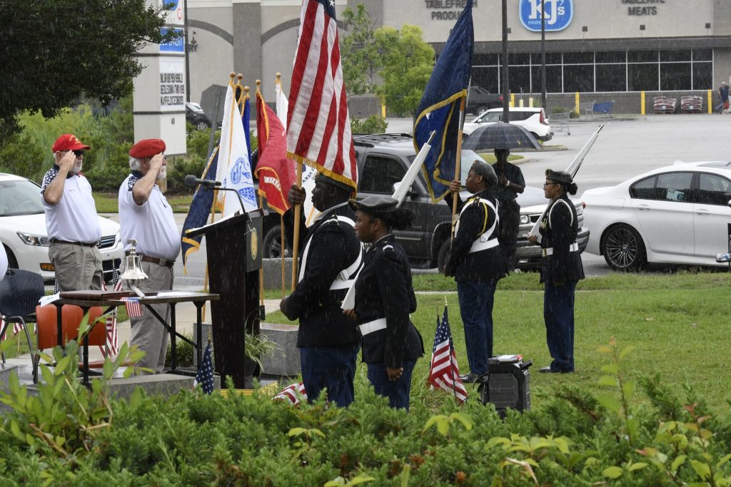 Military honor guard stands in formation holding flags during an outdoor ceremony, with a person saluting nearby. Small American flags are placed in the grass.