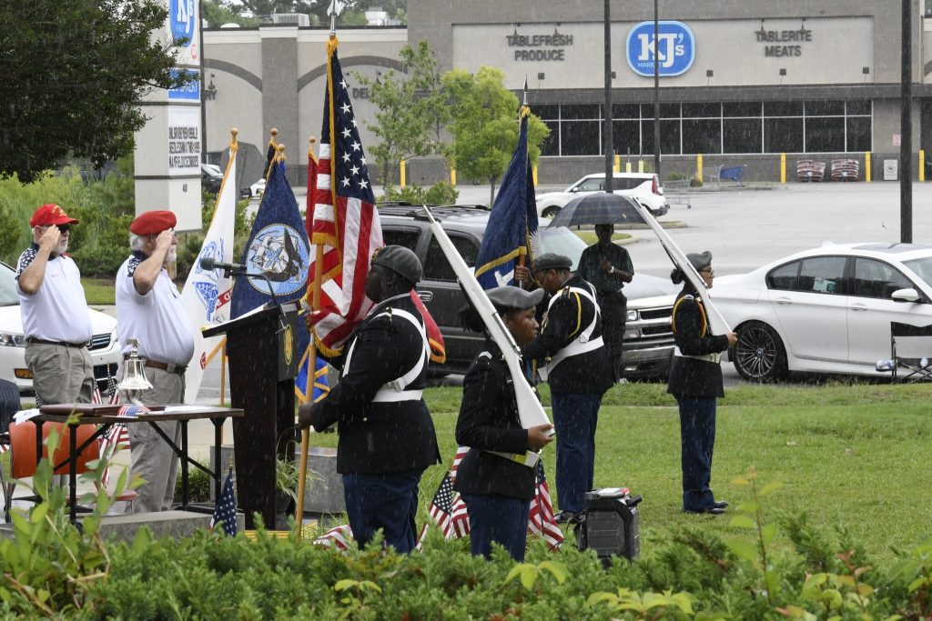 Uniformed honor guard holding flags during an outdoor ceremony, with American flags and onlookers in the background, near a parking lot and commercial buildings.