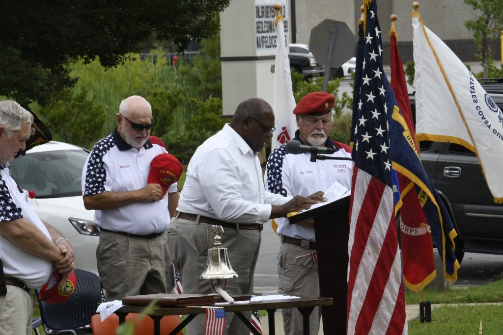 Three men participate in a patriotic outdoor ceremony, standing near flags and a podium, with one man holding his hat and another reading from a document.