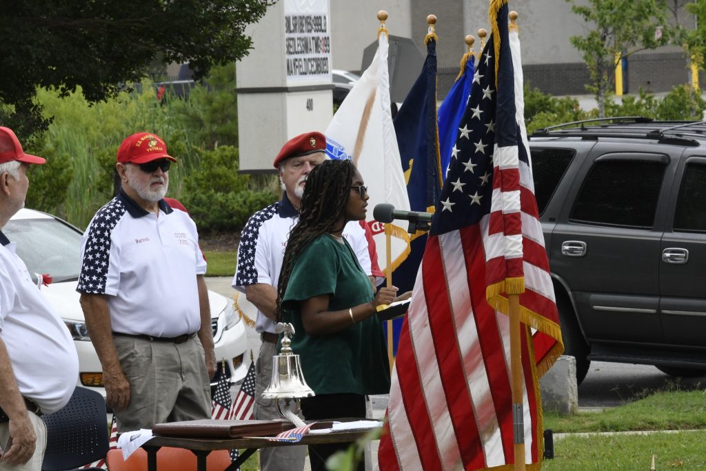 A woman speaks at an outdoor event with several flags, including the American flag, and three people in patriotic clothing standing nearby.