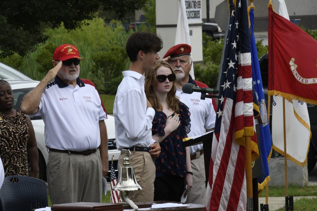 People stand near American flags, some saluting and others with hands on hearts, during a ceremonial outdoor event.