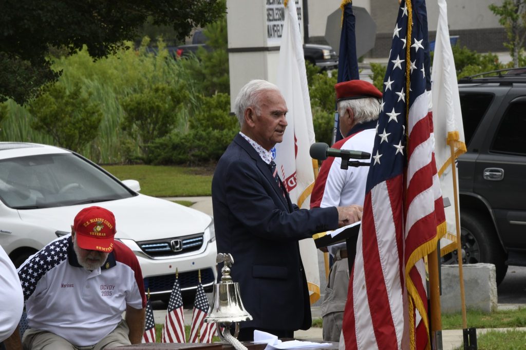 A man speaks at an outdoor event with American flags and a microphone, while others in uniform sit nearby. A car and greenery are in the background.