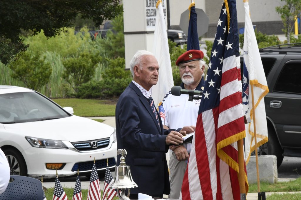 elderly man speaks at an outdoor event, standing beside a man in uniform and several American flags, with a car and greenery in the background.