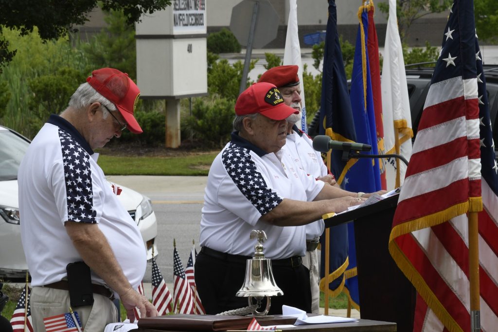 Three people in patriotic shirts and red caps participate in an outdoor ceremony with flags and small American flags in the background.