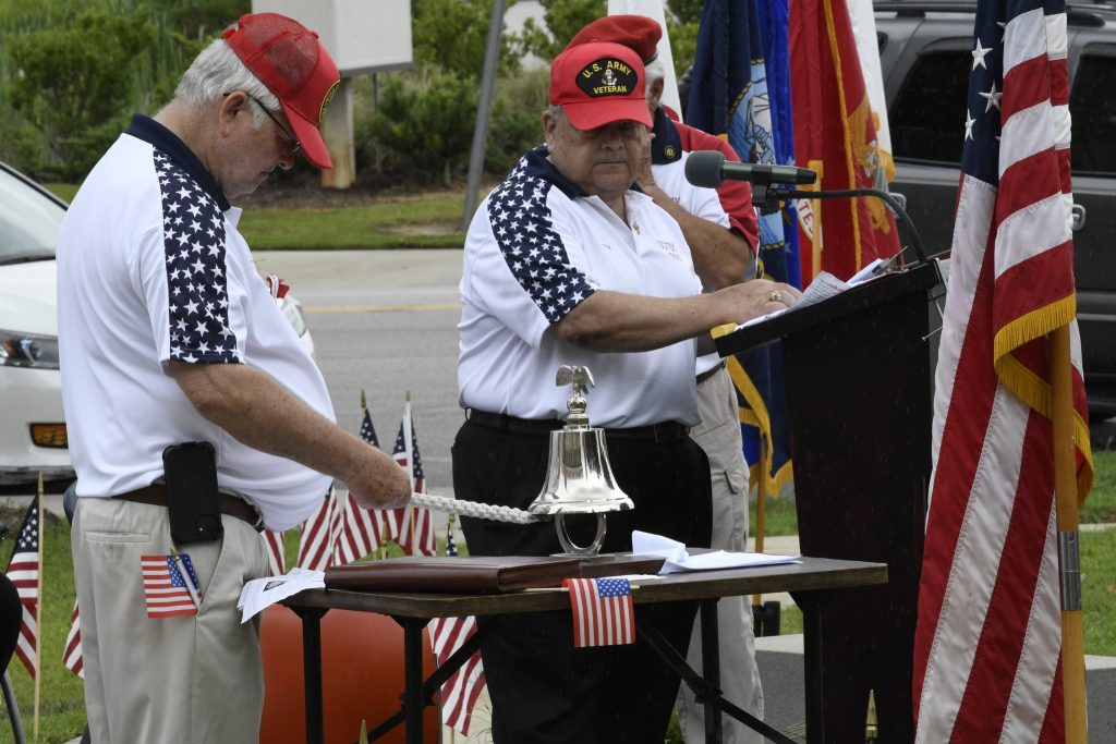 Two men in patriotic shirts and red caps participate in a ceremony with an American flag, bell, and microphone, standing beside a table with small flags.