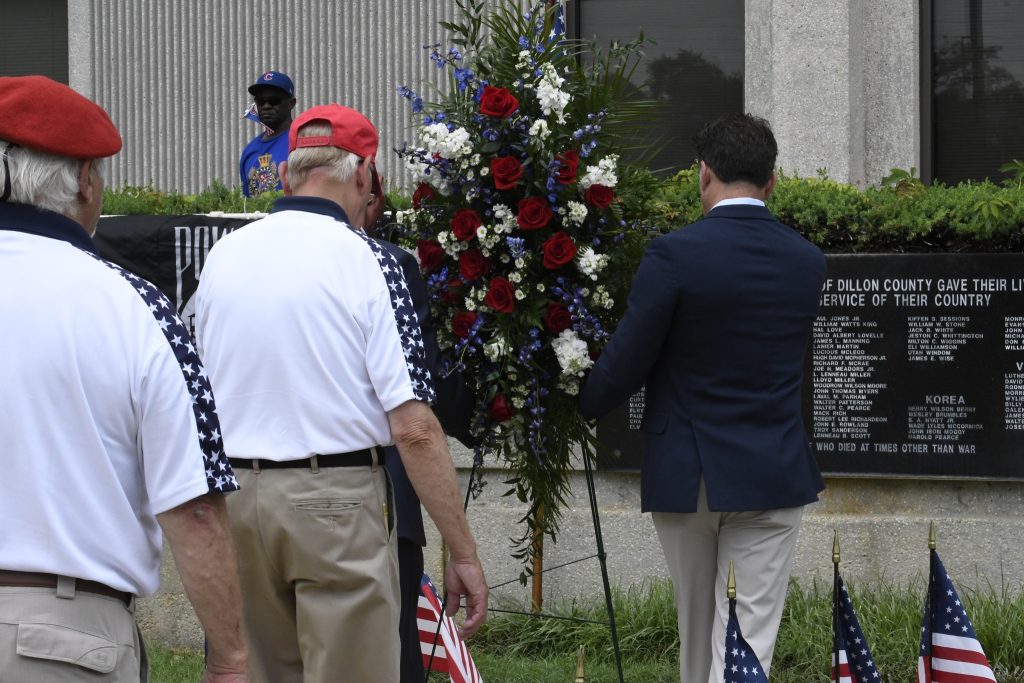 People stand near a red, white, and blue floral wreath outside, possibly during a memorial or commemorative event.