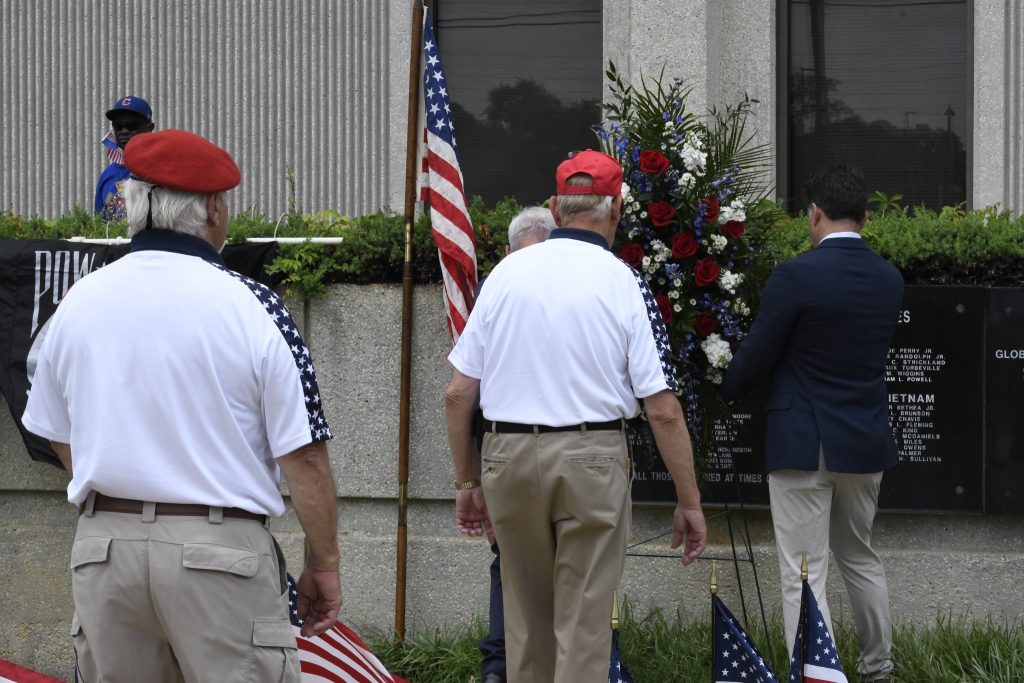 Men wearing red caps and white shirts stand near American flags and a floral wreath during an outdoor ceremony.