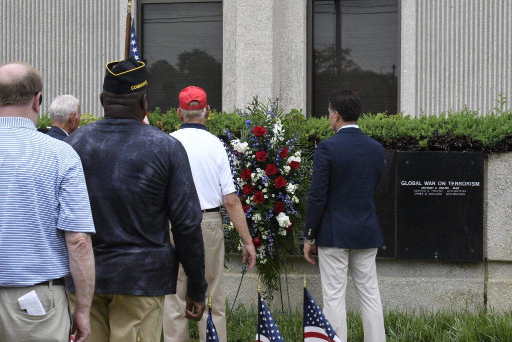 Three men stand in front of a building with American flags and a red, white, and blue floral wreath, possibly at a memorial or remembrance event.