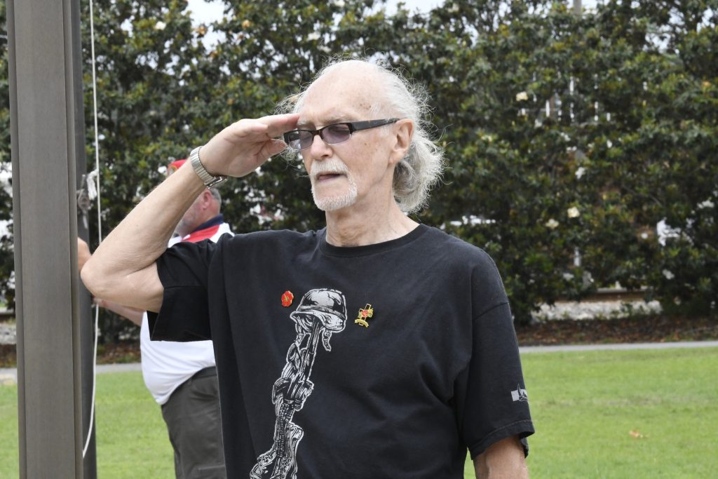 older man with gray hair and glasses is standing outdoors, saluting, and wearing a black t-shirt with a graphic design.