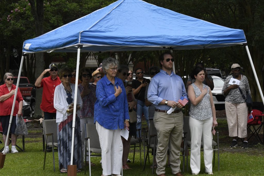 A group of people standing under a blue canopy outdoors, with hands over their hearts, participating in a ceremony or event.