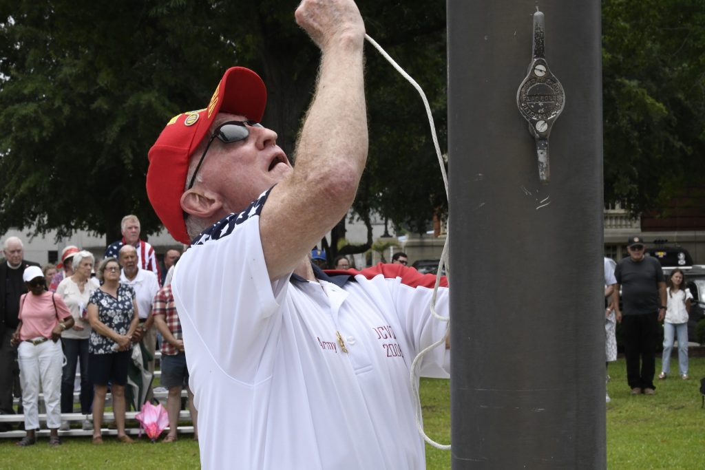 Man in a white shirt and red cap raising a flag on a flagpole during an outdoor event.
