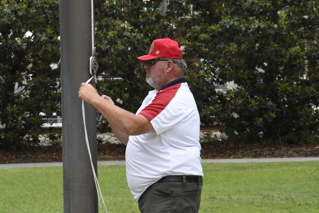 Man in a red cap and white shirt tying or adjusting a rope on a flagpole outdoors.