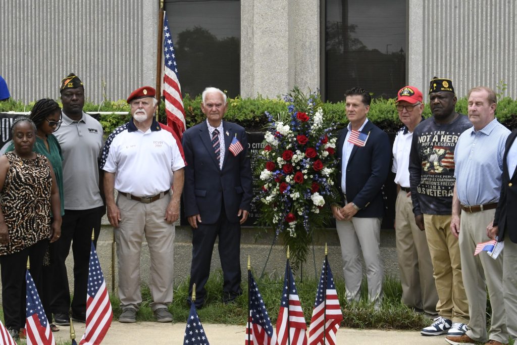 Five men stand by an American flag and a red, white, and blue wreath, with small U.S. flags on the ground in front of them.