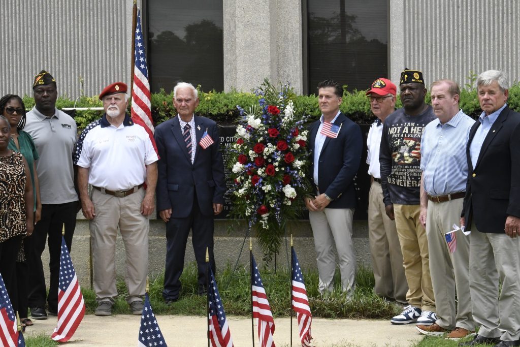 A group of men stand in front of a memorial wreath with American flags, posing for a photo outside a building.