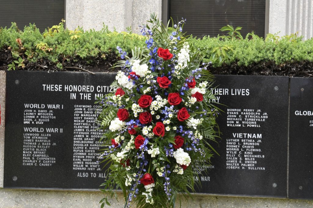 A memorial wall with engraved names, and a large wreath of red, white, and blue flowers placed in front, honoring fallen individuals.